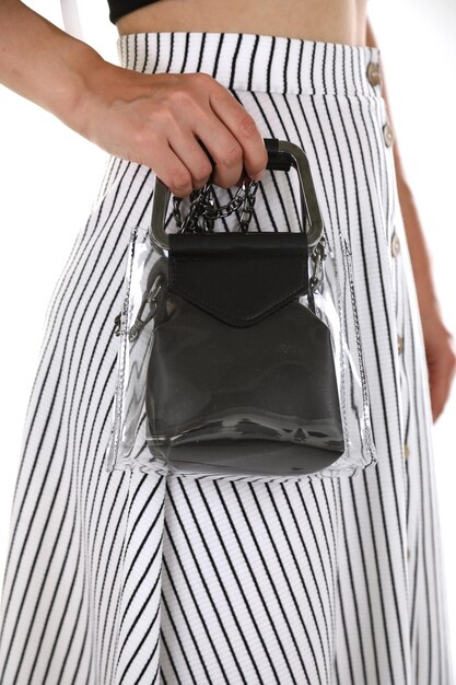 A stylish woman with a monochromatic outfit carrying a clear, transparent handbag filled with colorful accessories like silk scarves, sunglasses, and lipstick tubes. The background is a minimalist white studio, putting all the attention on the handbag and its playful contents.