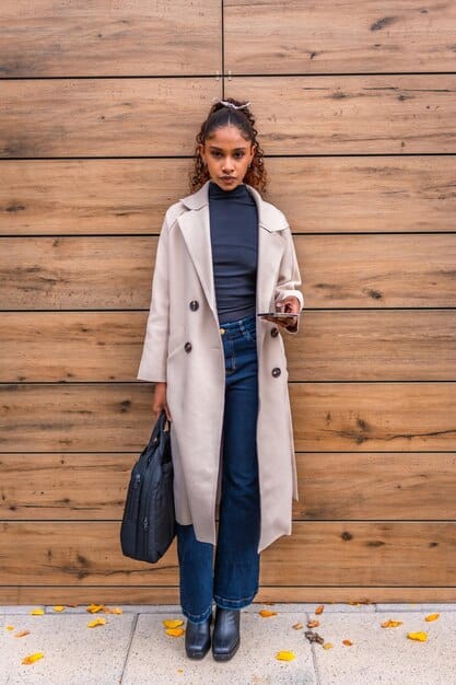 A celebrity (e.g., Zendaya) wearing a beige trench coat over a black turtleneck and wide-leg trousers. She is accessorized with sunglasses and ankle boots. The setting is a city street, capturing a candid and stylish moment.