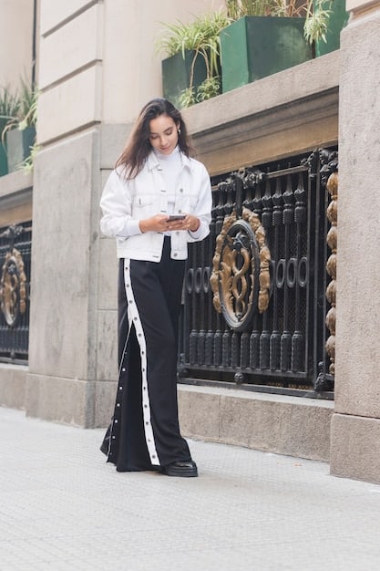 A celebrity (e.g., Meghan Markle) wearing high-waisted black tailored trousers, a white blouse, and pointed-toe heels. She is carrying a structured handbag and wearing minimal jewelry. The setting is a professional or business-casual environment.