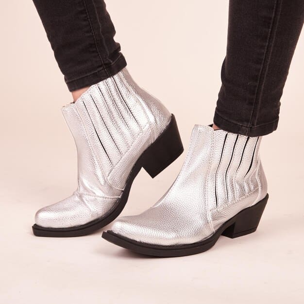 A close-up shot of a pair of silver metallic ankle boots with a chunky heel, placed on a reflective surface. The boots have intricate detailing, showcasing their futuristic design and high-end craftsmanship. The lighting highlights the shimmering texture of the metallic material.